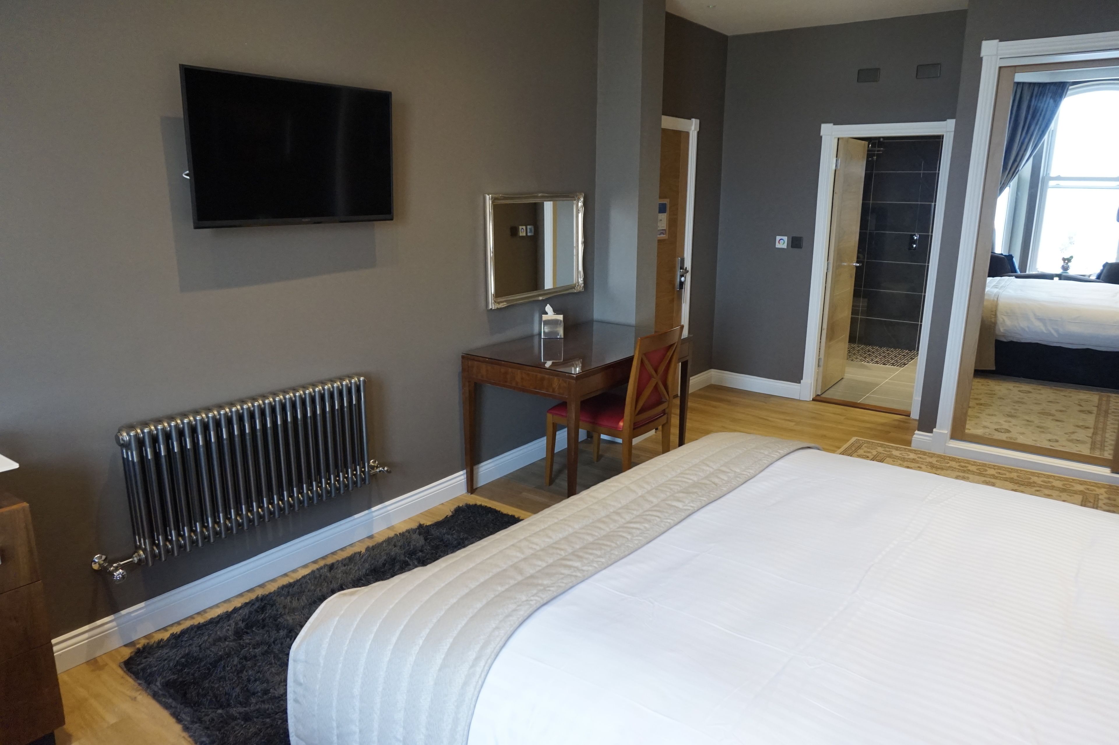 A hotel room with a bed, a desk with a chair, a TV, a mirror, a radiator, and a sliding door leading to a bathroom.