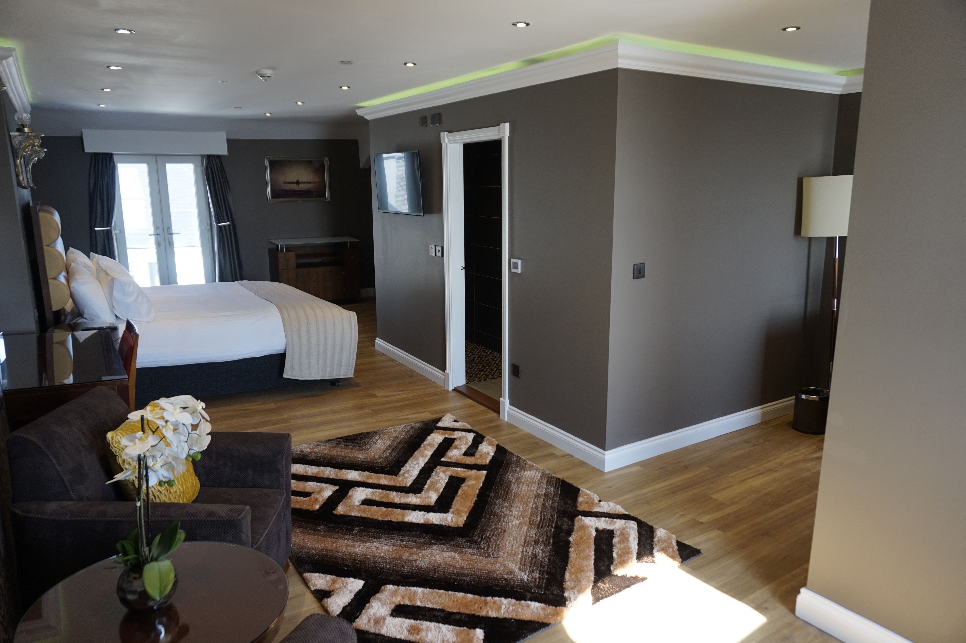 A modern hotel room with a large bed, a sofa, a dining table with a vase of flowers, a patterned rug, and a TV mounted on the wall.