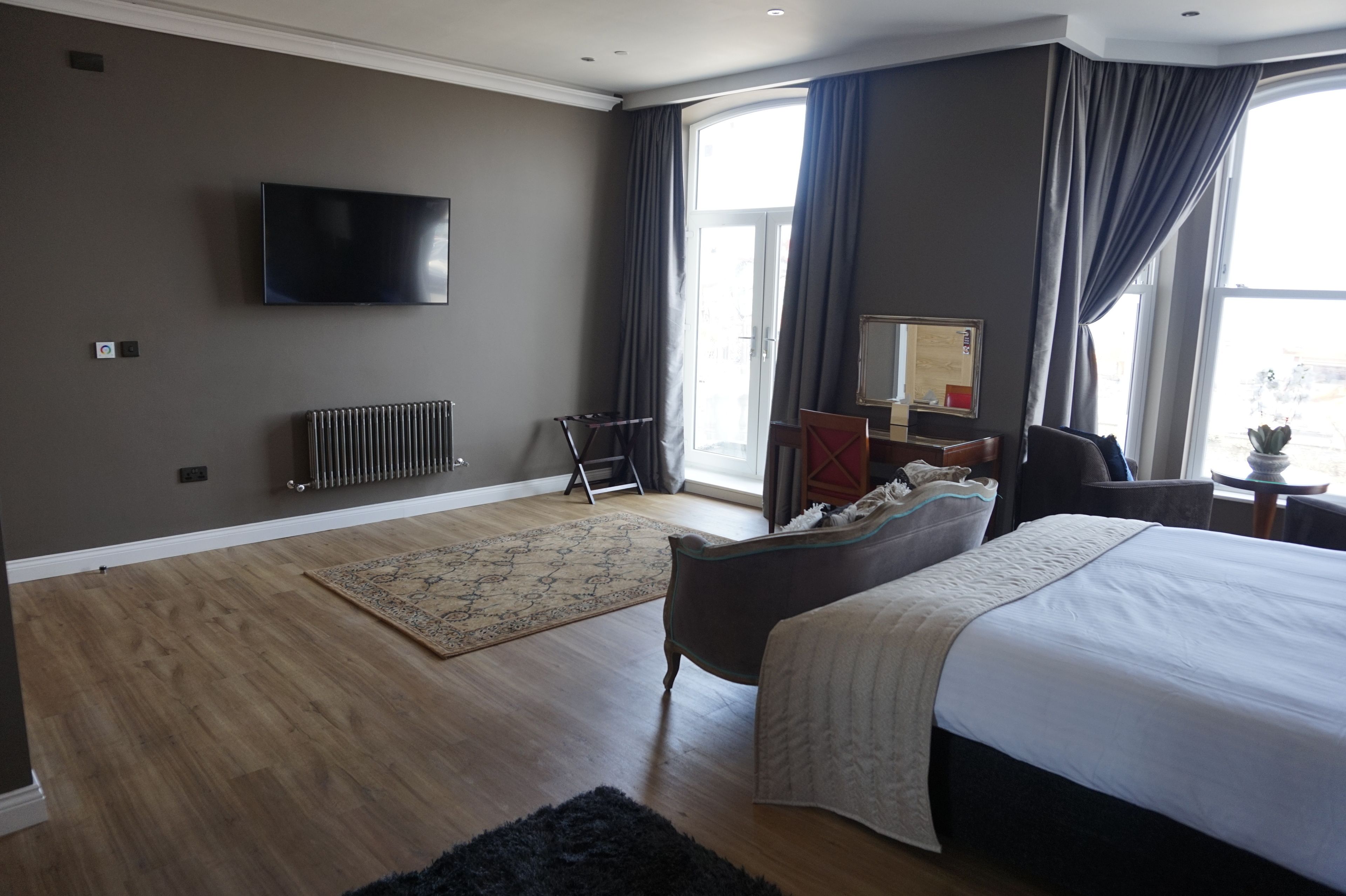 A modern hotel room with a large bed, a flat-screen TV mounted on the wall, a radiator, and a large window with curtains.
