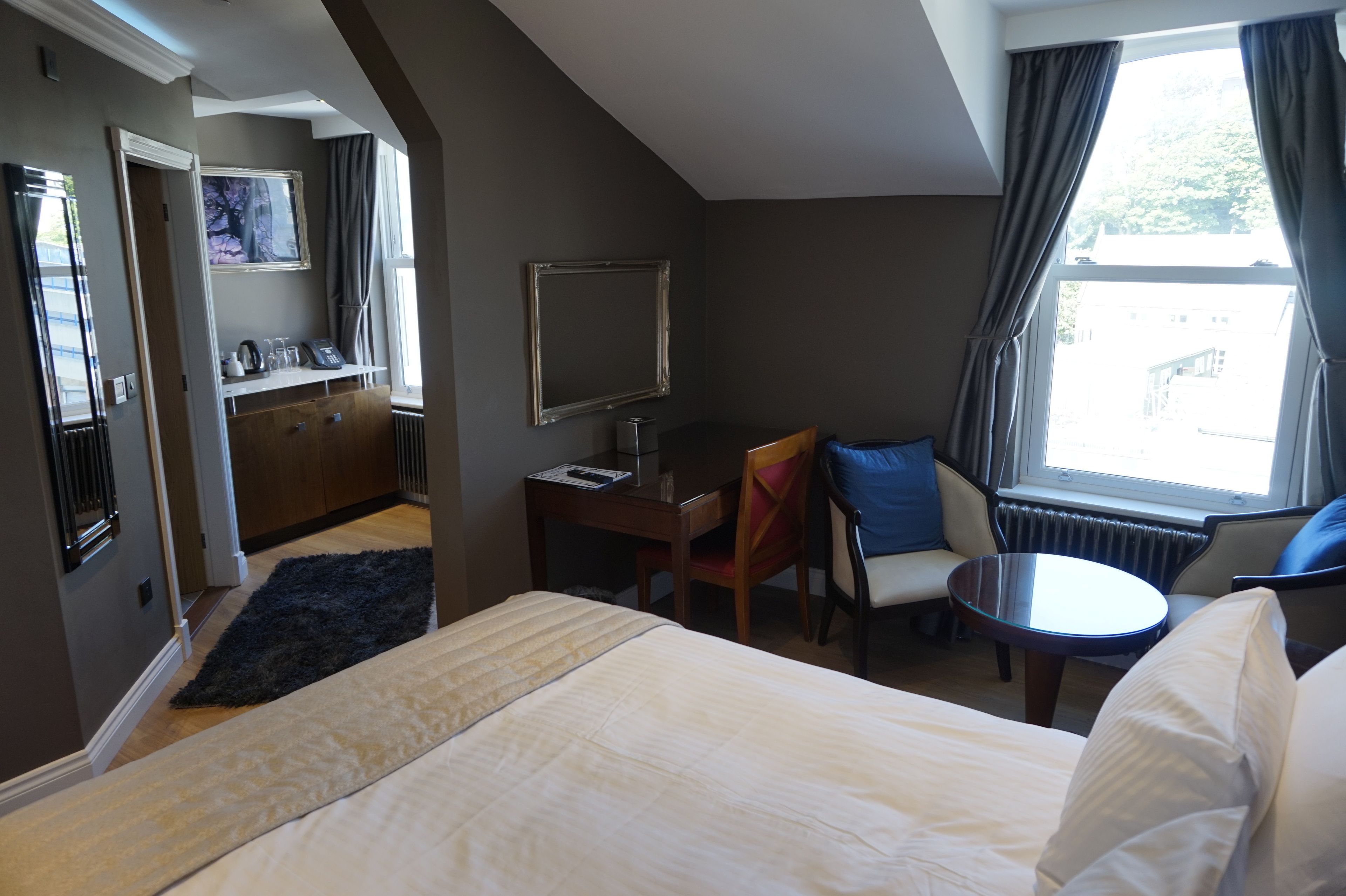 A hotel room with a bed, a desk, a chair, a small table, a TV, and a window with curtains.