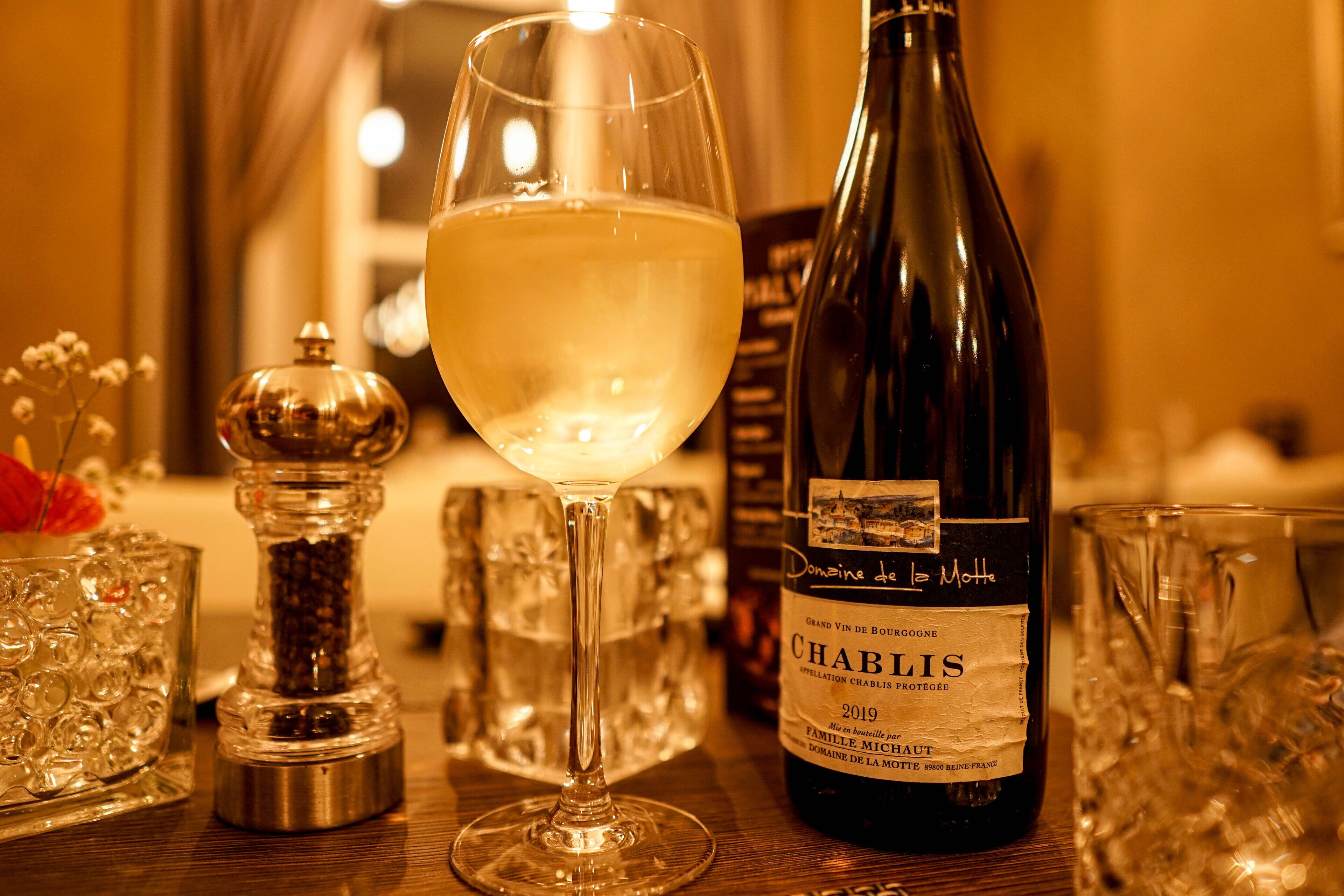 Una botella de vino Chablis 2019 Domaine de la Motte, una copa de vino blanco y un salero y pimentero sobre una mesa de madera.