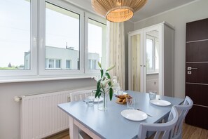 Un comedor con una mesa dispuesta para dos personas, sillas blancas, un florero con flores y vistas al exterior a través de amplias ventanas.