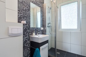 Un baño moderno con pared de diseño en blanco y negro, un lavamanos blanco con espejo arriba, ducha con puerta de cristal y una ventana con motivo de hojas.