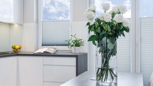 Una cocina moderna con armarios blancos, un florero transparente con rosas blancas, un cuenco de fruta y una ventana con persianas.