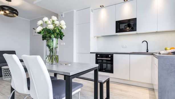 Una cocina moderna con armarios blancos, horno empotrado y microondas negro. Hay una mesa de comedor con dos sillas y un jarrón de flores.