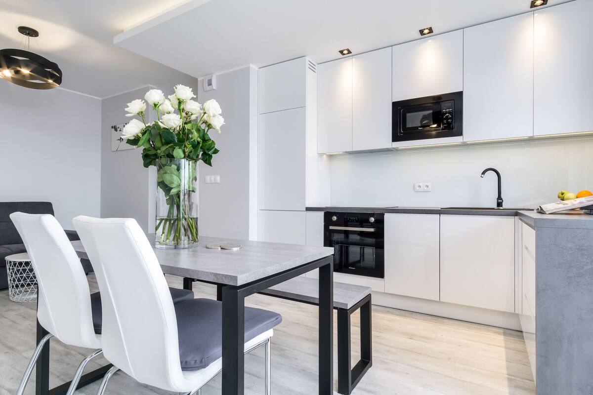 Une cuisine moderne avec des meubles blancs, un four encastré et un micro-ondes noir. On y trouve une table à manger avec deux chaises et un vase de fleurs.