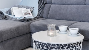 Un sofá gris con una revista y un libro, una mesa blanca con un tarro de vidrio y dos tazas, y una manta azul.