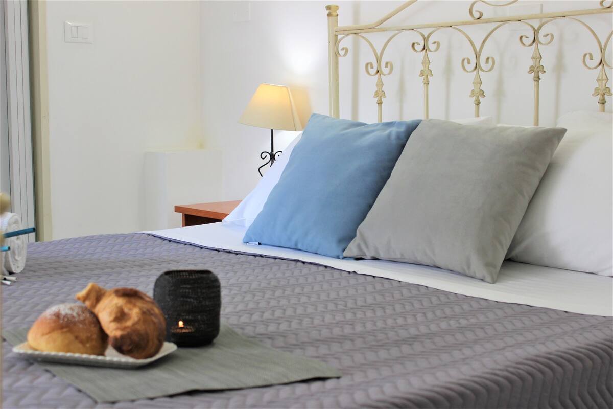 A neatly made bed with a patterned bedspread, two pillows, a bedside lamp, and a tray with two pastries.