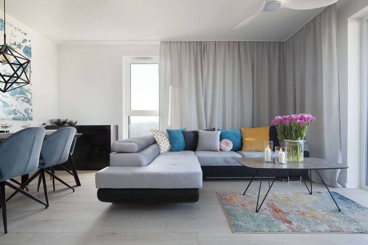 A modern living room with a sectional sofa, a dining area with chairs, a coffee table, and a large window with curtains.