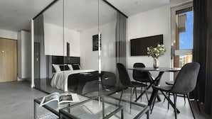 A modern hotel room with a glass partition separating the bedroom from the living area. There is a bed with white bedding, a glass coffee table, and a dining table with chairs. A flat-screen TV is mounted on the wall, and a vase with flowers is on the table. - Elite Apartments Cityview Center (Gdansk)