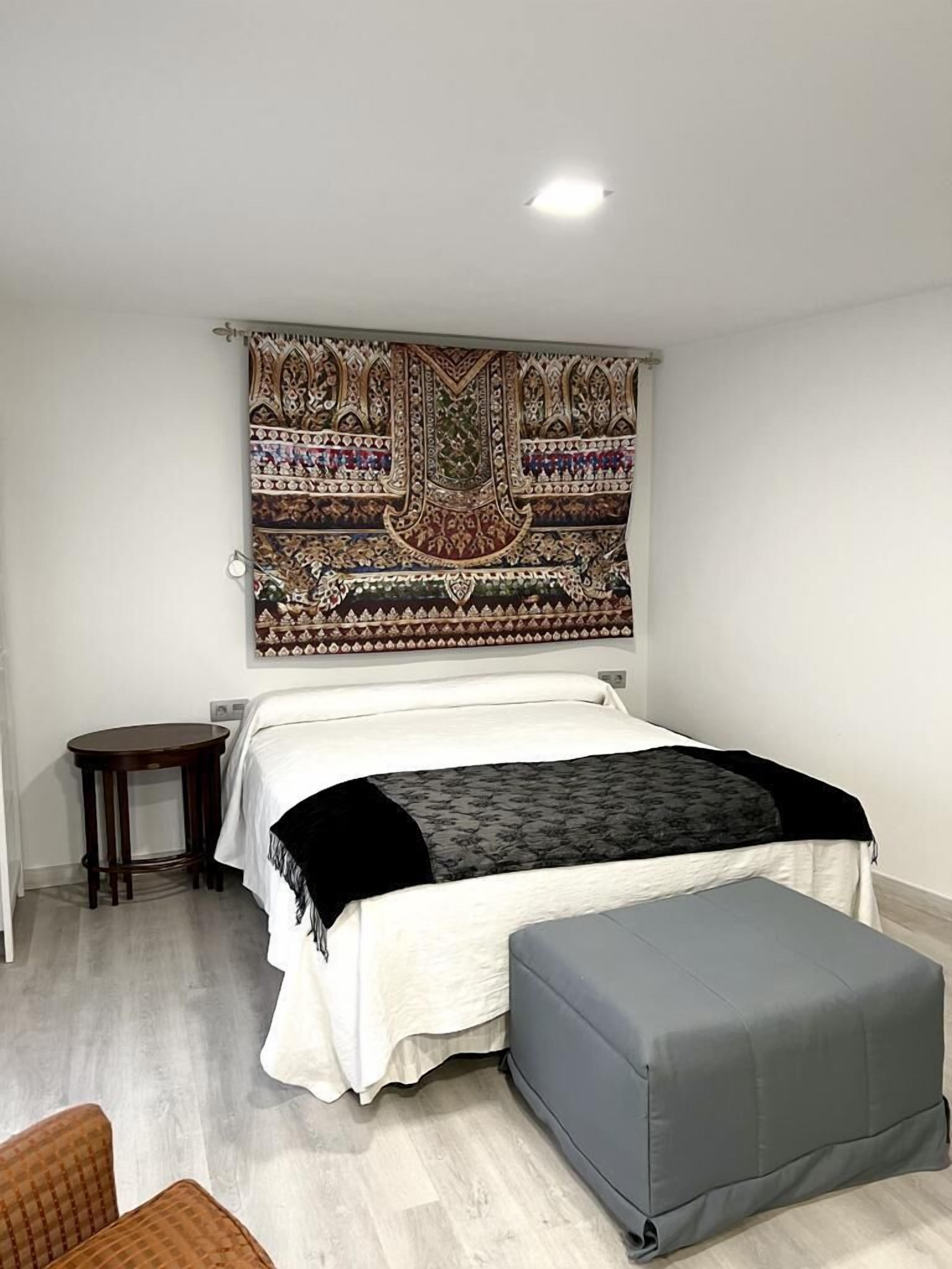 A bedroom with a bed, a bedside table, a chair, and a tapestry on the wall.