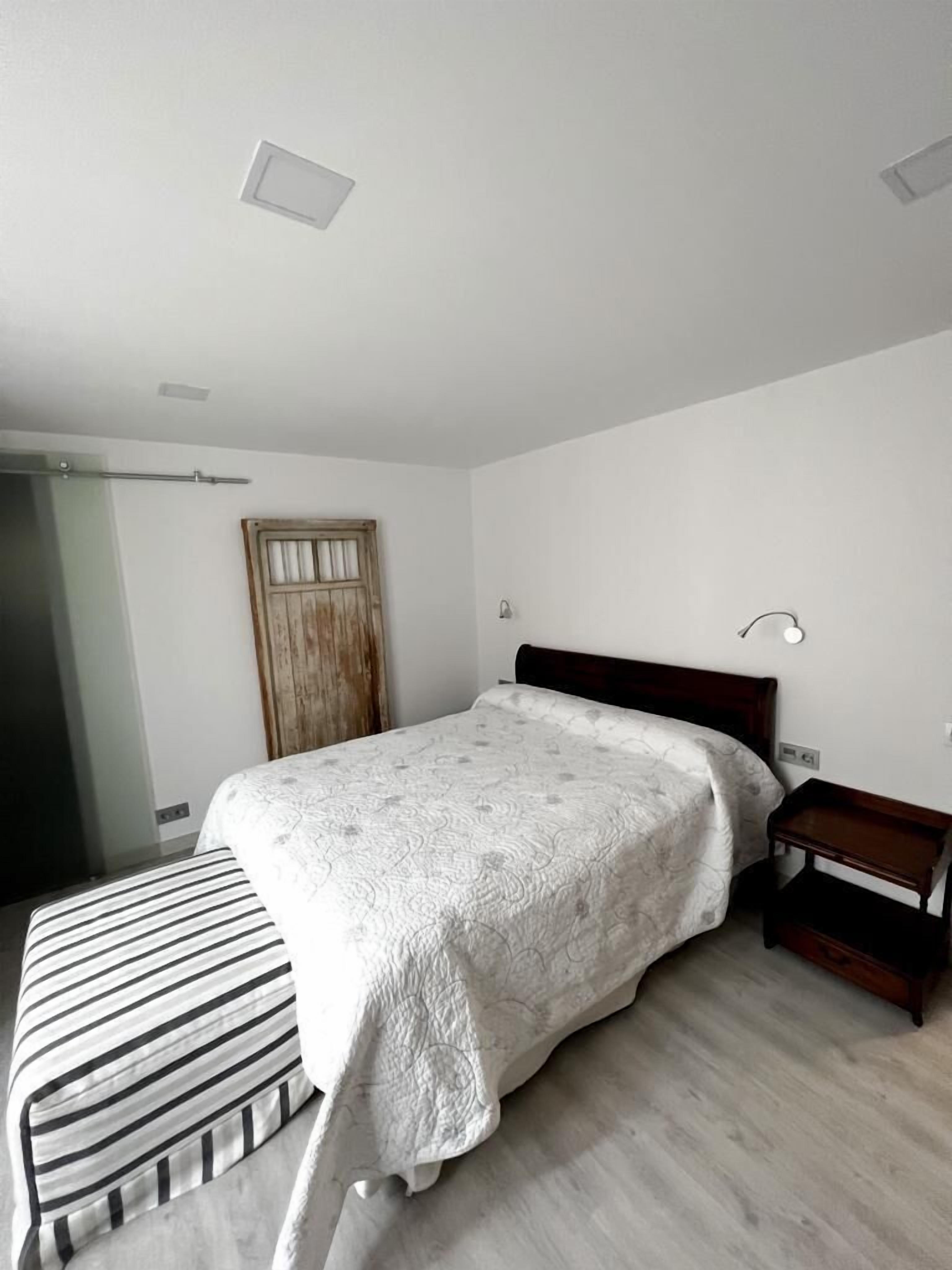 A bedroom with a bed, a bedside table, a wooden door, and a light fixture.