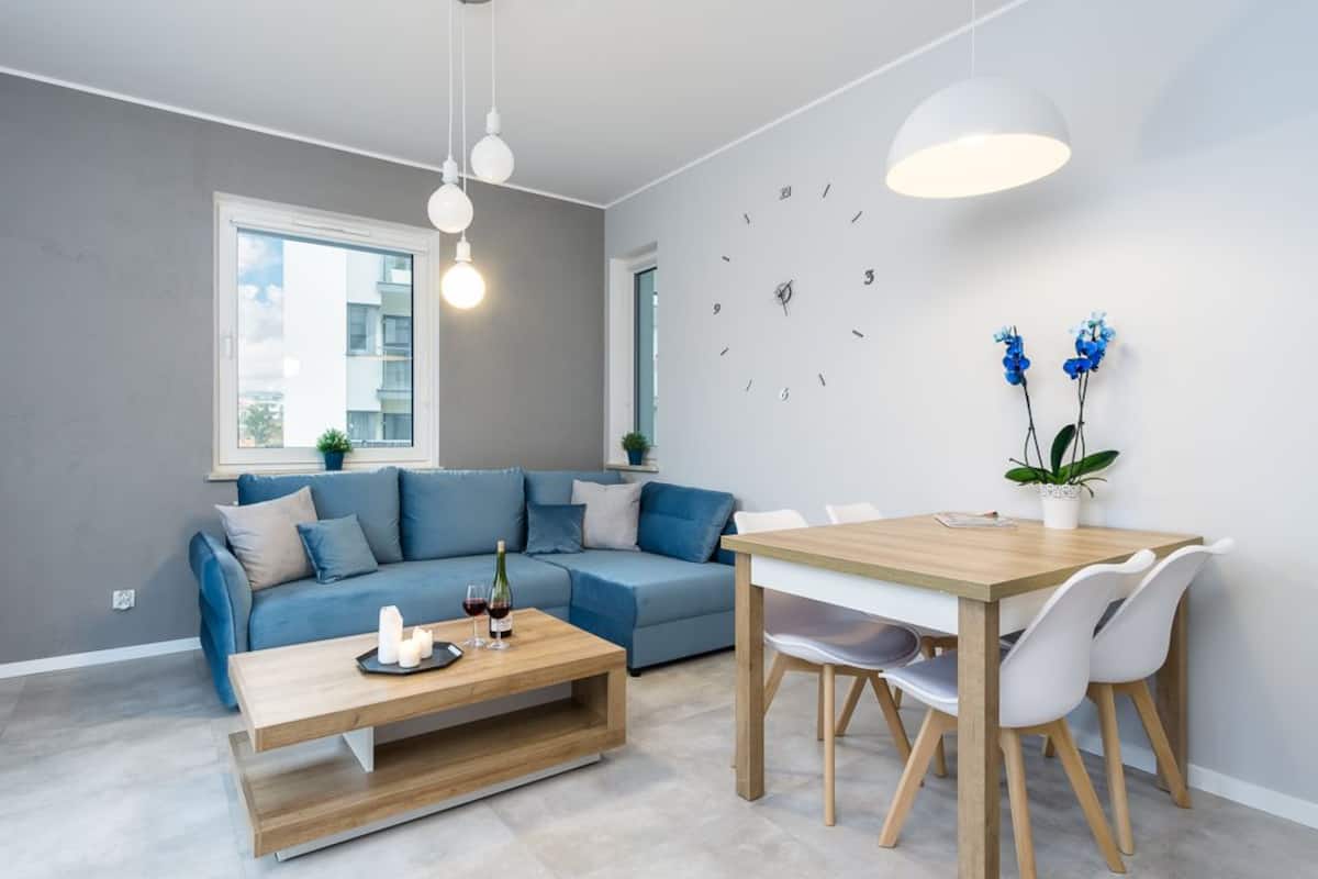 A modern living room with a blue sofa, a wooden coffee table, and a dining table with chairs. There is a wall clock and a pendant light above the dining table.