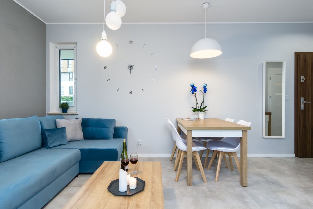 A modern living room with a blue sofa, a wooden dining table with chairs, a coffee table with candles and wine glasses, and pendant lighting.