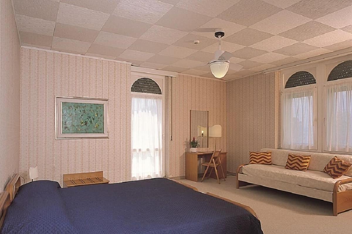 A hotel room with a large bed, a desk with a chair, a sofa, and a ceiling fan.
