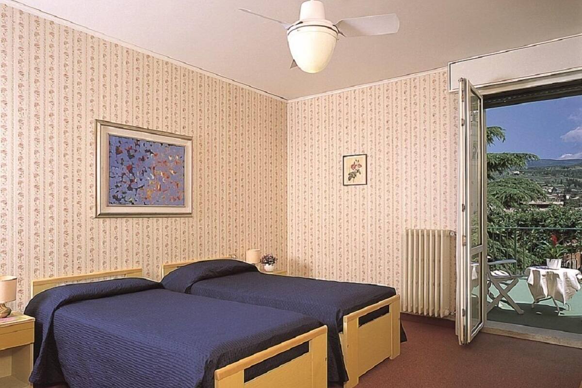 A bedroom with two beds, a ceiling fan, a radiator, and a balcony with a view.