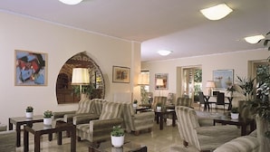 Un vestíbulo amplio con varias zonas de estar, incluyendo sofás y sillones, dispuestos alrededor de una mesa de centro. Hay varias plantas en macetas y obras de arte enmarcadas en las paredes.