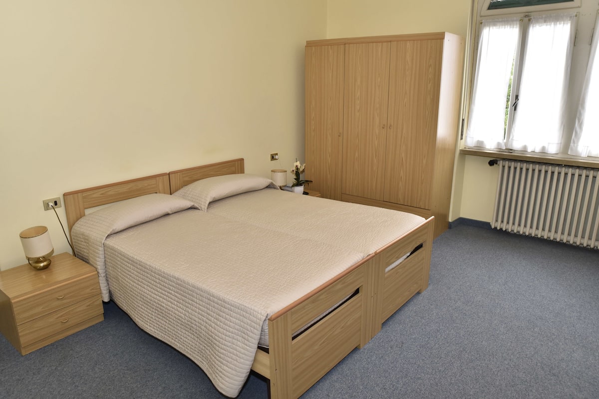 A bedroom with a bed, wooden wardrobe, bedside table, lamp, and a window with curtains.