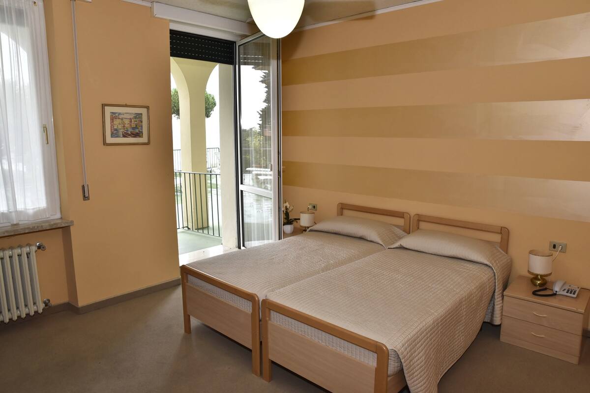 A bedroom with two beds, a radiator, a bedside table with a lamp, and a window with curtains.