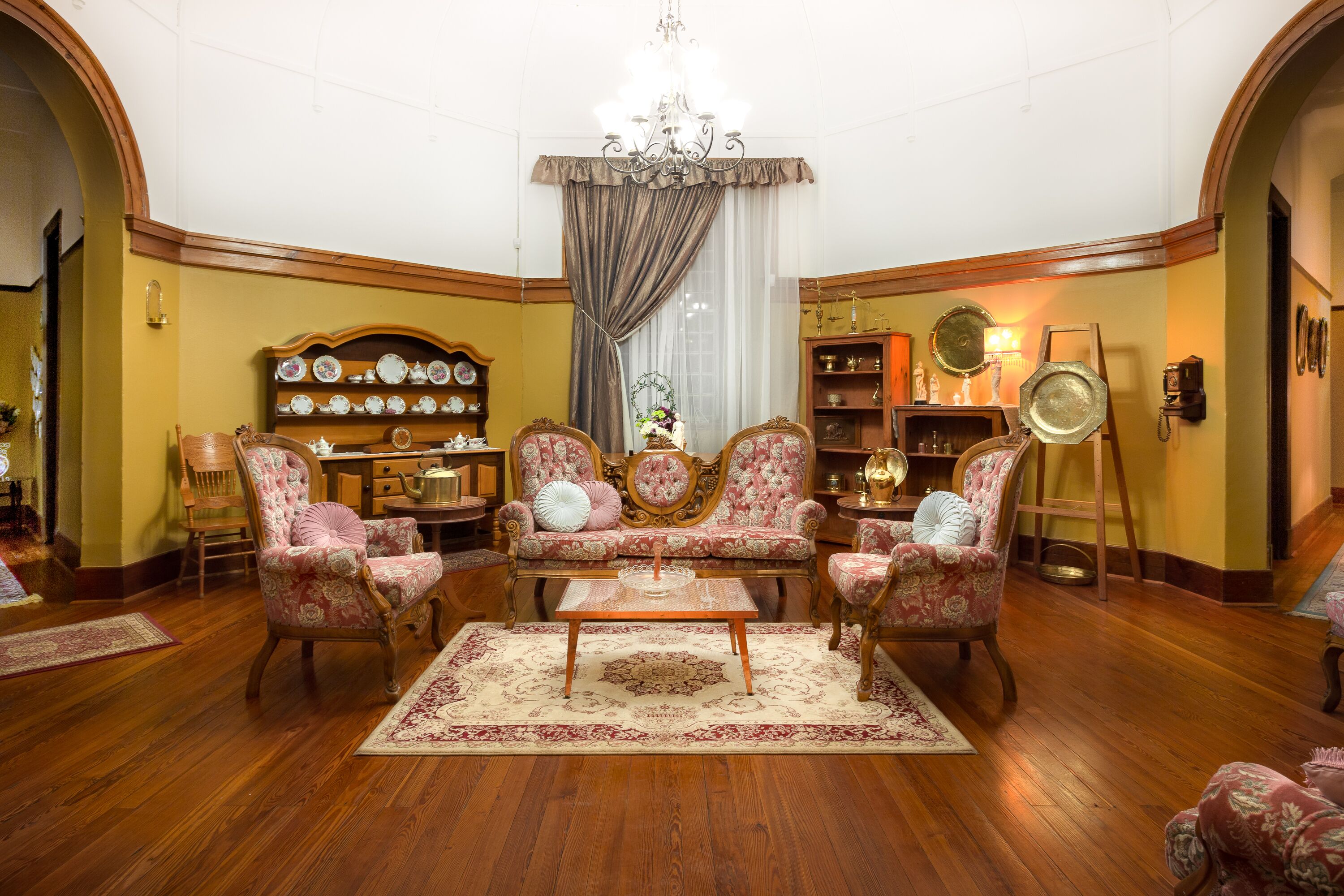 Een klassieke woonkamer met een houten vloer, een centrale salontafel, armstoelen met bloemenprint, een bank en een kroonluchter.