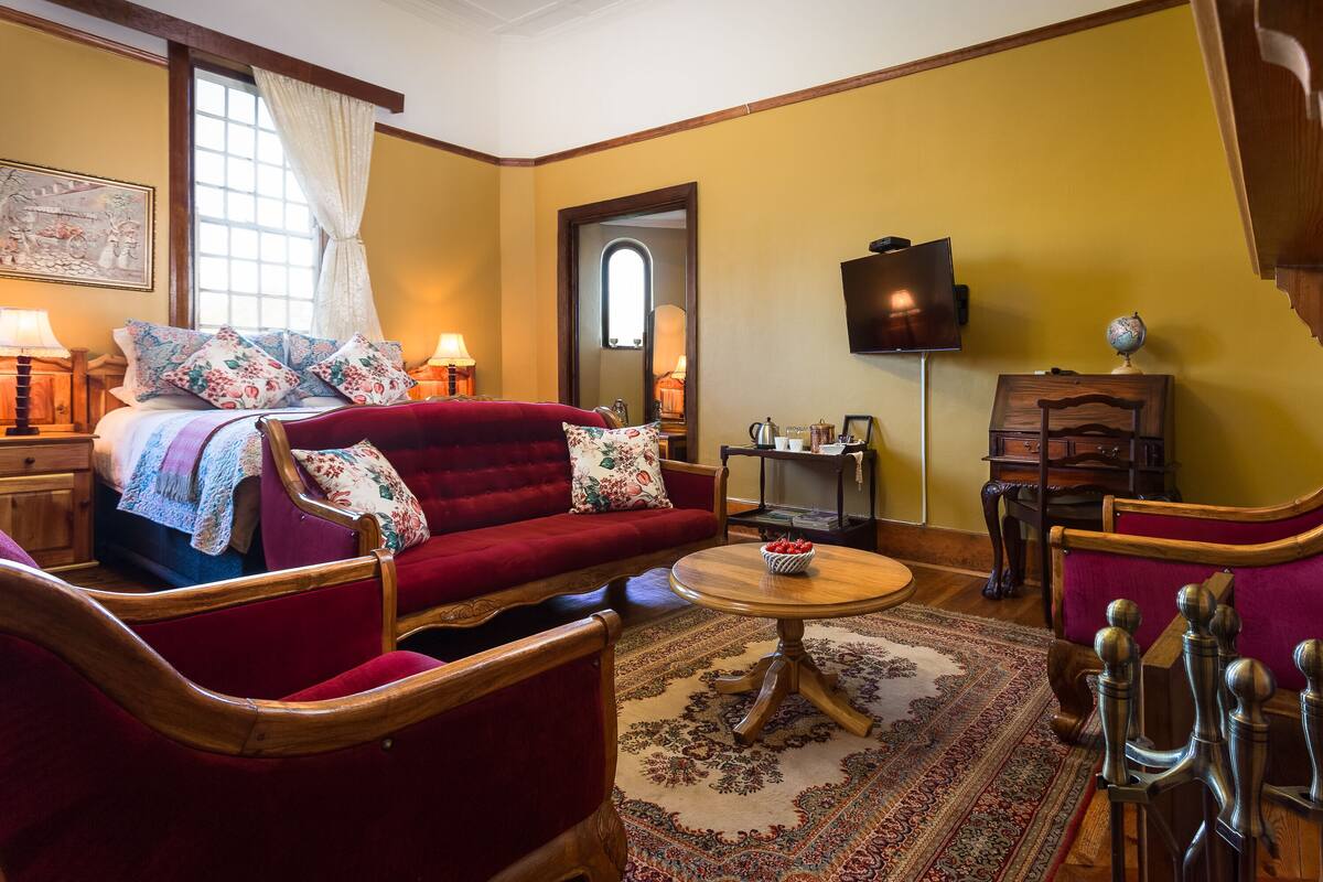 Un cuarto acogedor con un sofá rojo, sillones de madera, una mesa redonda de madera y una cama con almohadones florales.