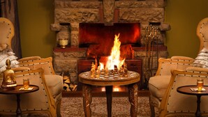 A cozy room with a fireplace, two armchairs, a round table with chess pieces, and a candle on the table.