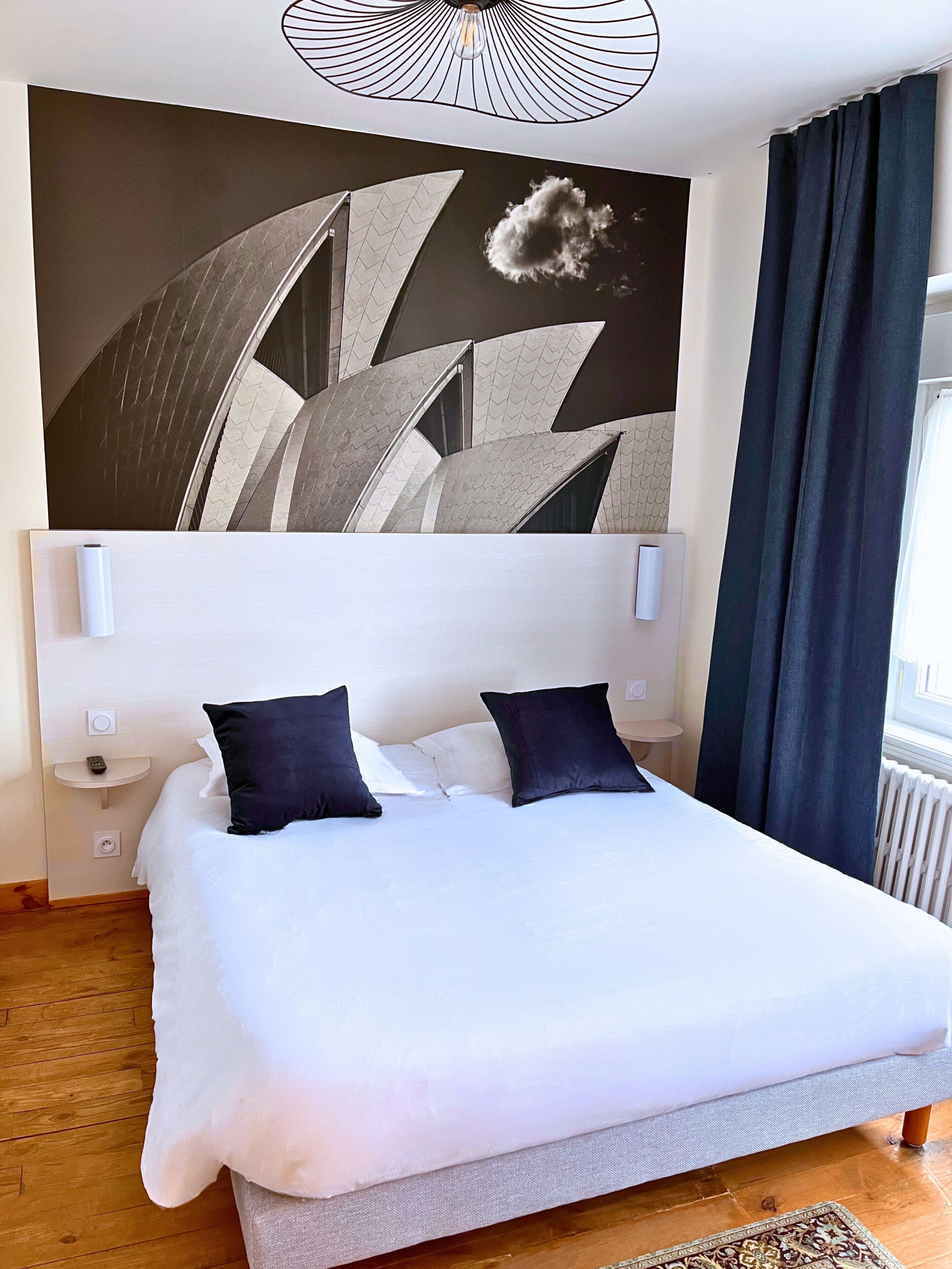 A modern hotel room with a large bed, a headboard with a geometric design, a wall-mounted light fixture, and a view of the outdoors.