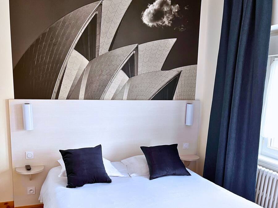 A modern hotel room with a large bed, a headboard with a geometric design, a wall-mounted light fixture, and a view of the outdoors.