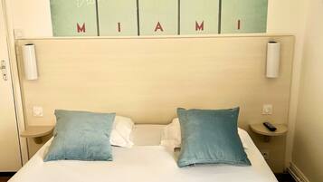 Une chambre d’hôtel avec un lit, deux oreillers, une table de chevet et un tableau représentant des palmiers accroché au mur, ainsi qu’une pancarte sur laquelle on peut lire : « Visitez MIAMI ».