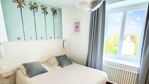 A bedroom with a large bed, a wall-mounted light fixture, a window with curtains, and a wall art piece. - Hôtel Restaurant La Maison (Trébeurden)