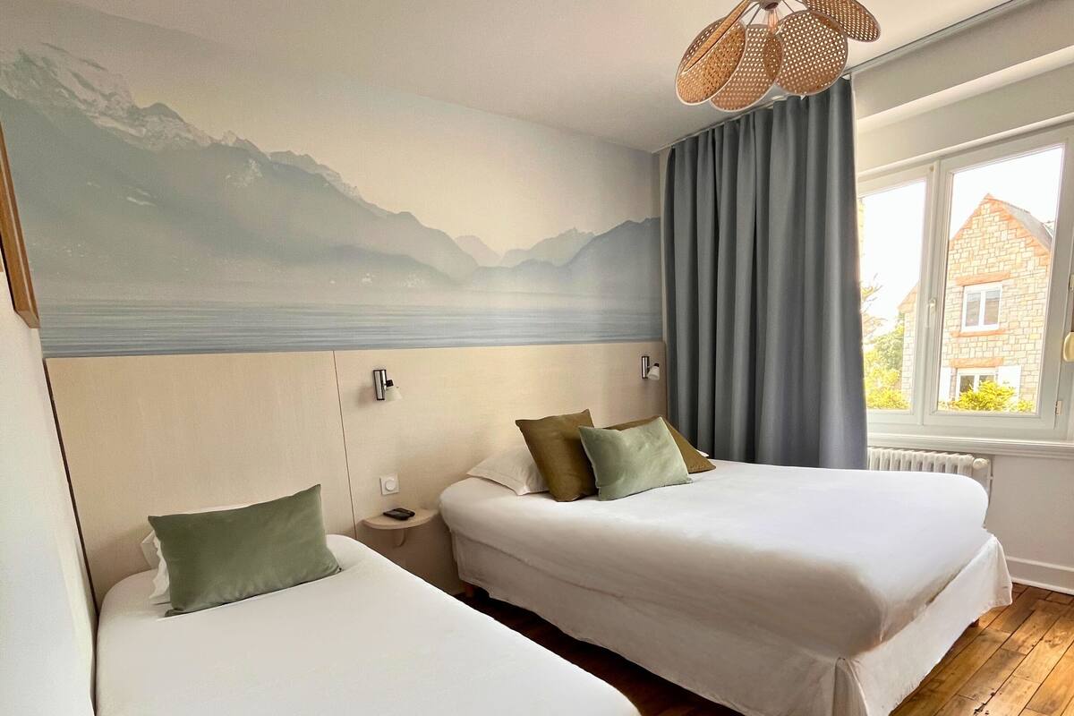 A hotel room with two beds, a large window with curtains, a wooden floor, and a wall mural of mountains.