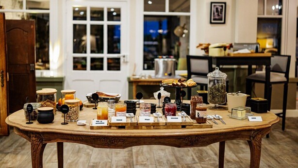 A wooden table with various jars and containers labeled with food items, set in a room with a wooden floor and white walls. - Hôtel Restaurant La Maison (Trébeurden)