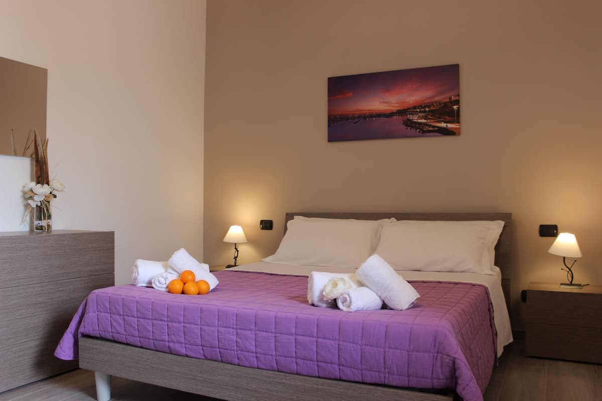 A hotel room with a bed, two bedside lamps, a nightstand, a wall-mounted painting, and a dresser with a vase of flowers.