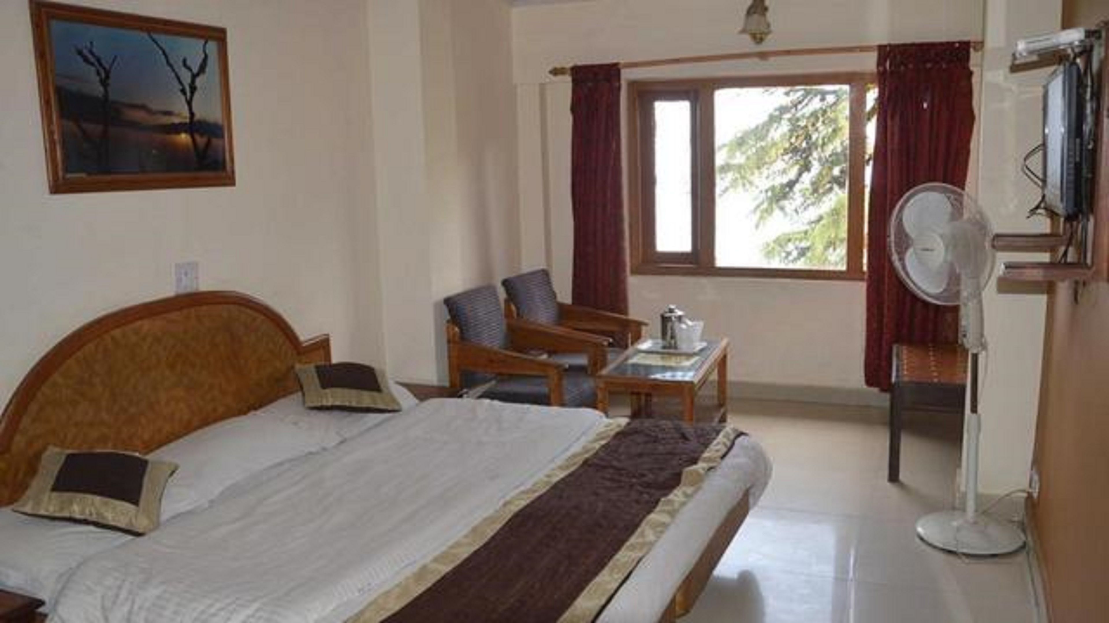 A hotel room with a bed, a chair, a small table, a fan, and a window with curtains.