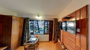 A compact room with a wooden wardrobe, a small table with a vase and bottles, a TV mounted on the wall, and a window with curtains. - Hotel Kahkashan (Dehradun)