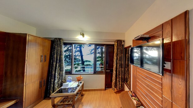 Una habitación compacta con un armario de madera, una mesita con un florero y botellas, un televisor montado en la pared y una ventana con cortinas.