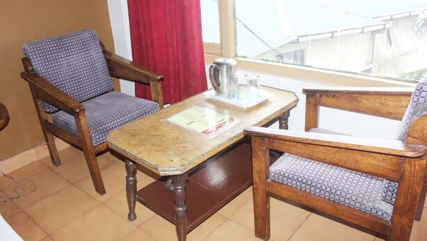 A room with two wooden chairs with patterned cushions, a small wooden table with a menu, and a window with a red curtain. - Hotel Kahkashan (Dehradun)