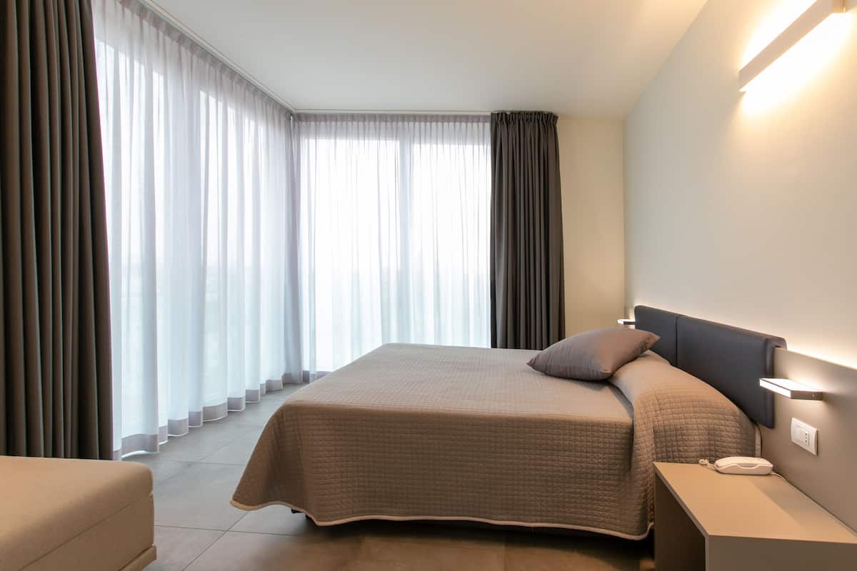 Ein Hotelzimmer mit einem großen Bett, einem Nachttisch mit Lampe und Blick durch durchsichtige Vorhänge nach draußen.
