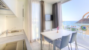 Una cocina moderna con un lavamanos y un comedor con mesa y sillas. Hay un televisor montado en la pared y vistas al mar a través de grandes ventanales.