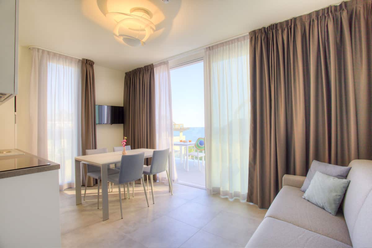 Ein modernes Wohnzimmer mit einer Couch, einem Esstisch und Stühlen. Es gibt eine Küchenzeile mit einer Arbeitsplatte sowie einen Fernseher. Das Zimmer verfügt über große Fenster mit Vorhängen.