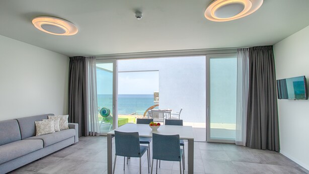 Una sala de estar moderna con sofá, mesa de comedor y sillas, un balcón con vista al mar y un televisor de pantalla plana montado en la pared.