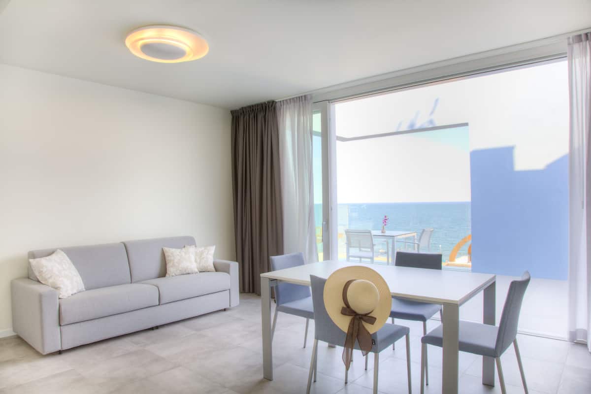 Ein modernes Wohnzimmer mit Sofa, Esstisch, Stühlen, einem großen Fenster mit Meerblick und einer Deckenleuchte.
