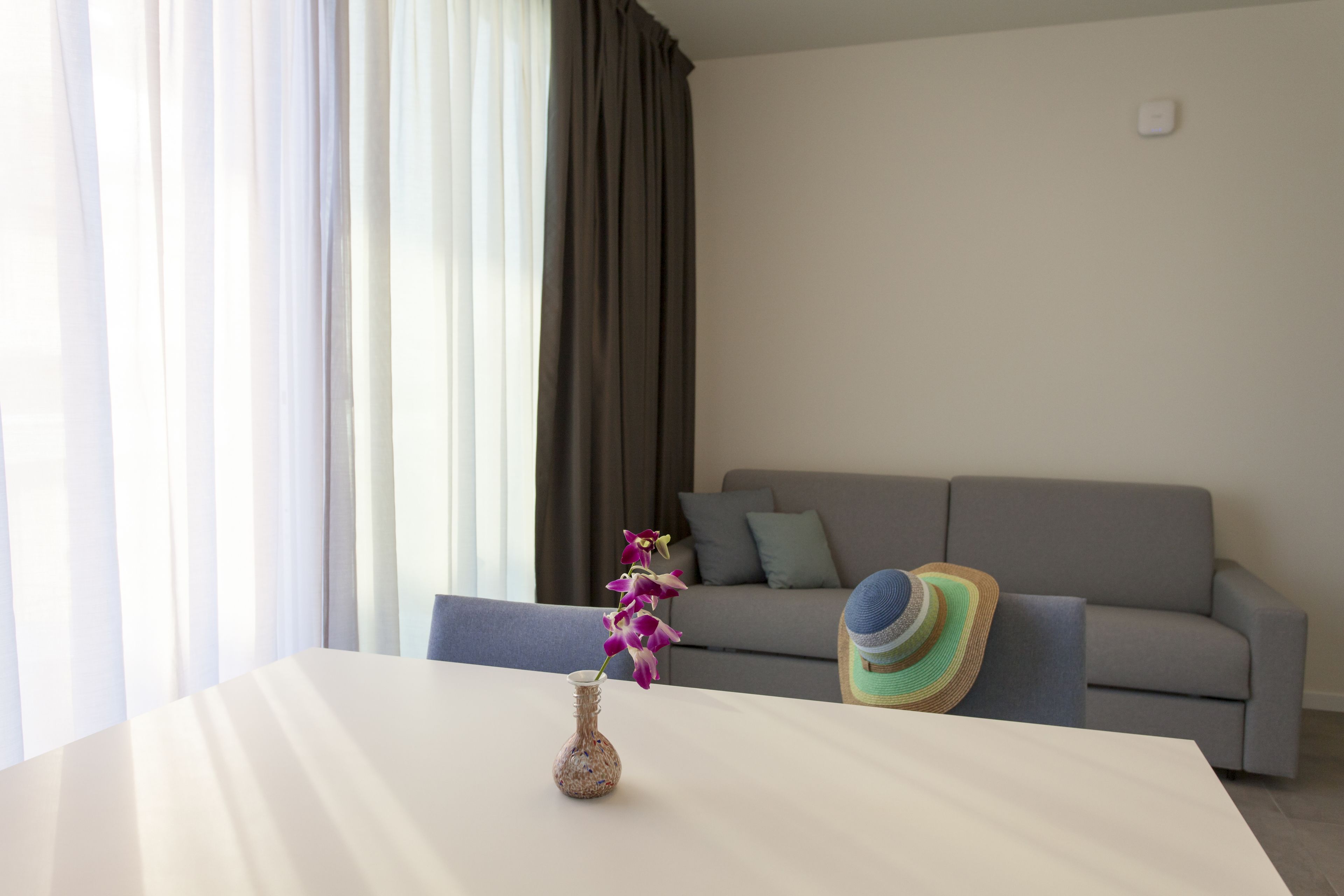 Una sala de estar moderna con una mesa blanca, un jarrón con flores, un sofá gris y un sombrero colorido sobre el sofá.