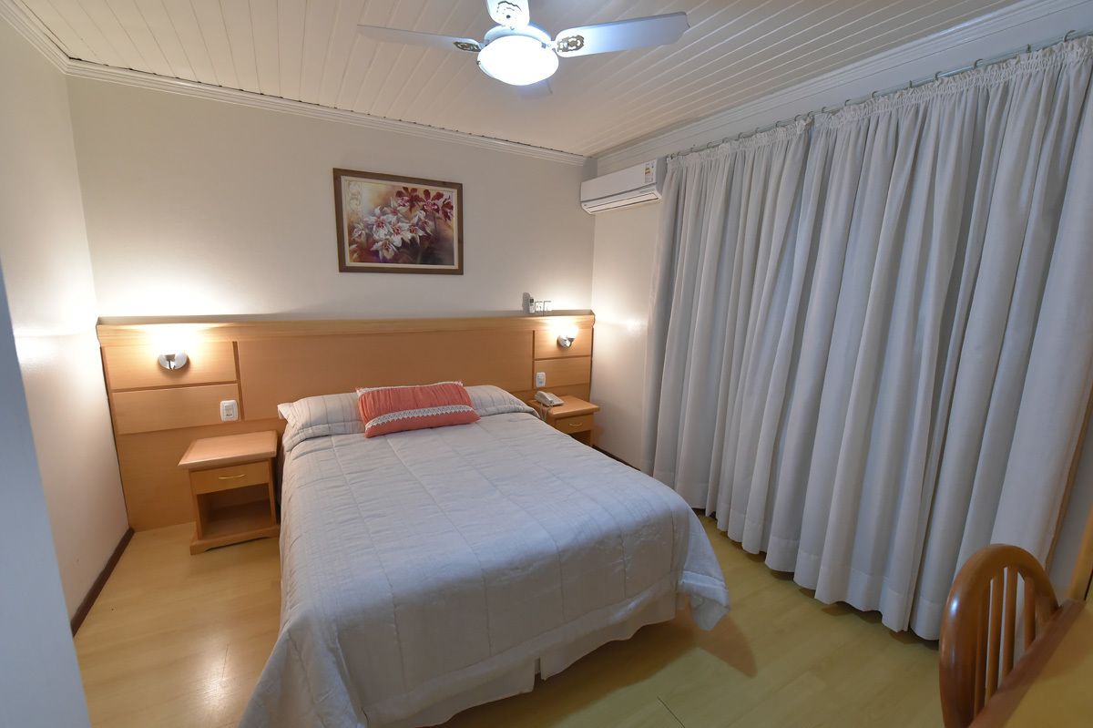 Habitación de hotel con cama, mesitas de noche, ventilador de techo, un cuadro y una ventana con cortinas.