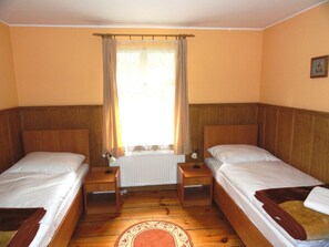 Una habitación con dos camas individuales, cabeceros de madera, un radiador, una mesita de noche con lámpara y una ventana con cortinas.