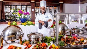 Montaje de bufé con diversos platos, un chef con uniforme y una zona de bar con botellas al fondo.