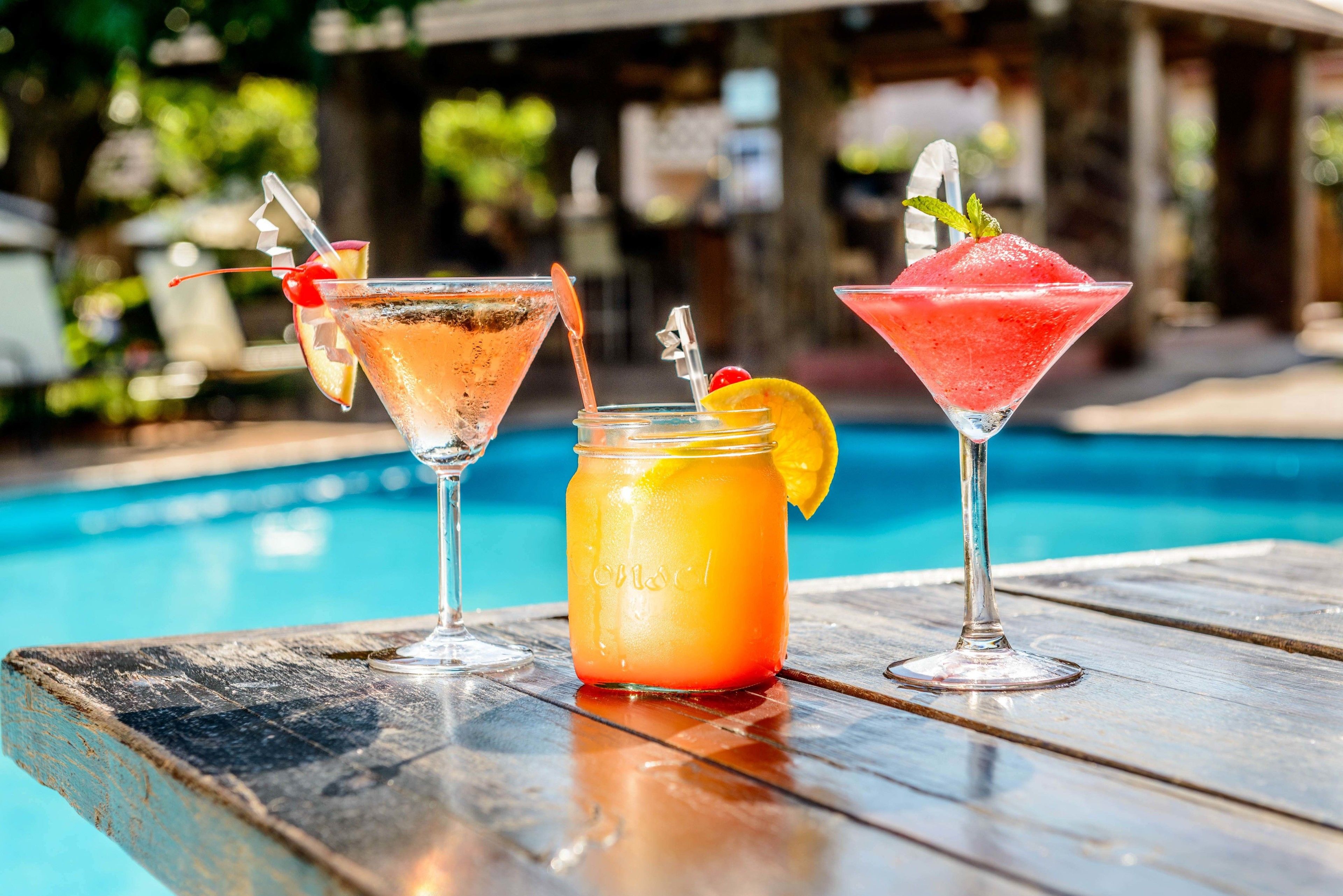 Three drinks on a wooden table by a pool.