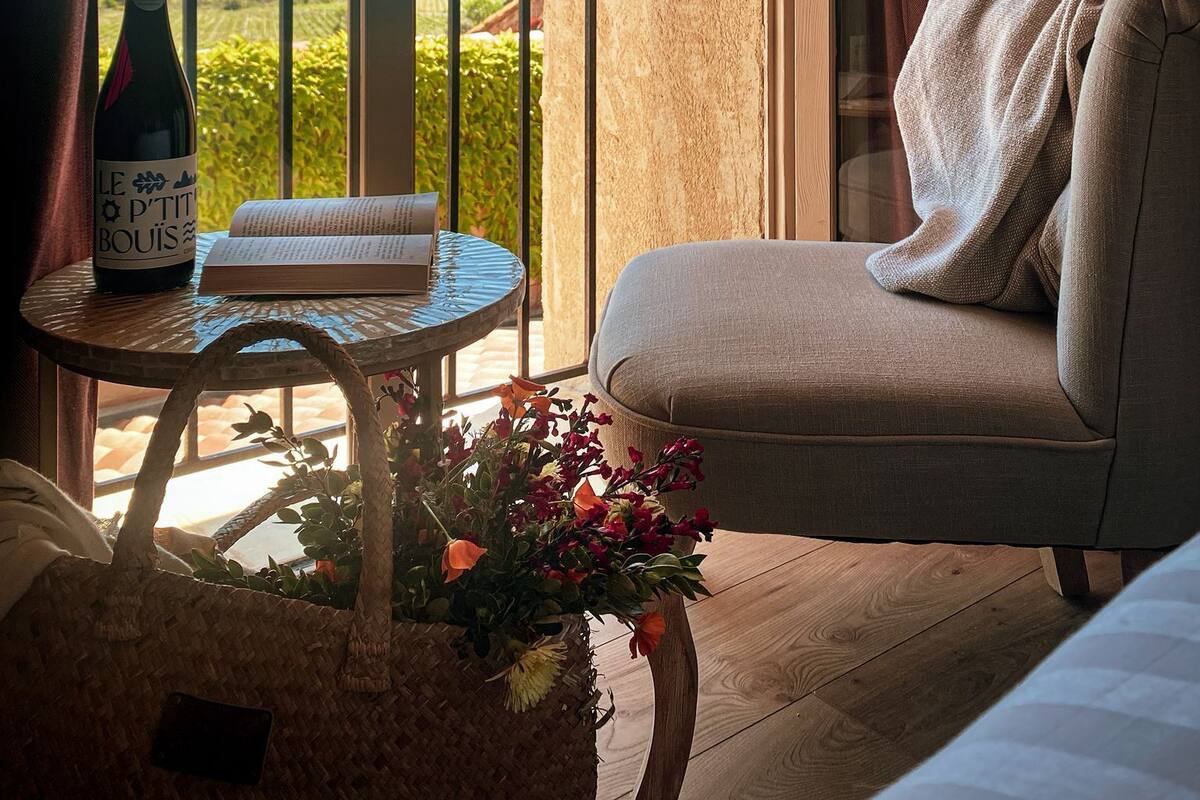 Un balcón con vistas a la vegetación, una botella de vino, un libro y una cesta de mimbre con flores.