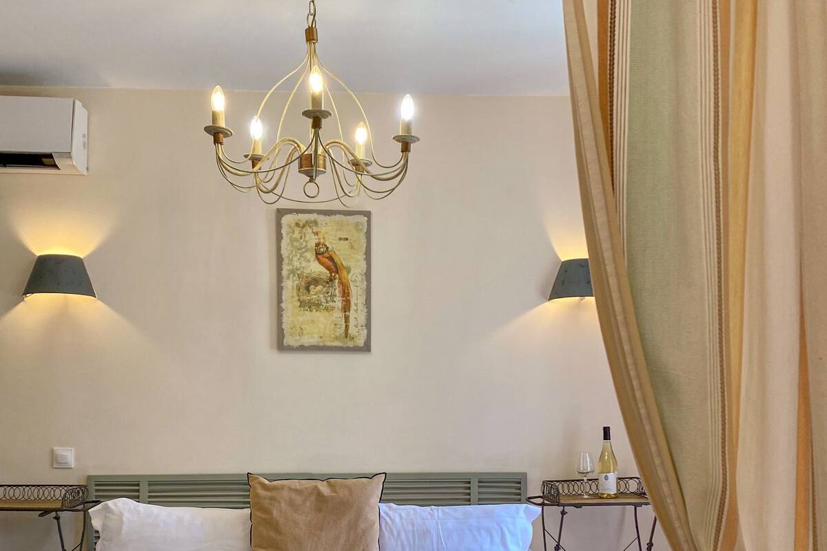 Un dormitorio con cama, candelabro, luces en la pared, un cuadro y una mesa con una botella y vasos.