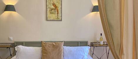Un dormitorio con cama, candelabro, luces en la pared, un cuadro y una mesa con una botella y vasos.